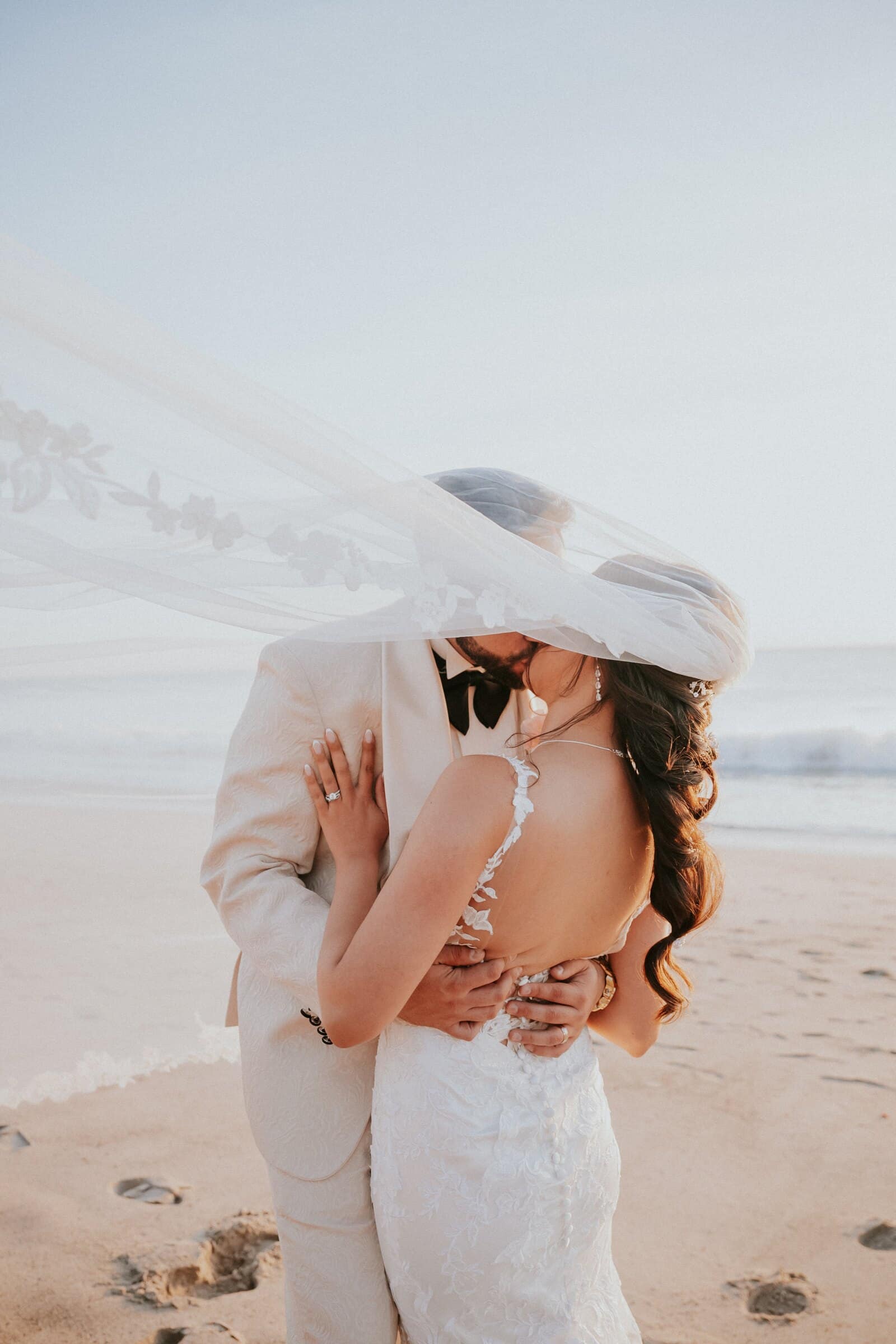 Ariana and Dom kissing on the beach as her veil lifts in the wind